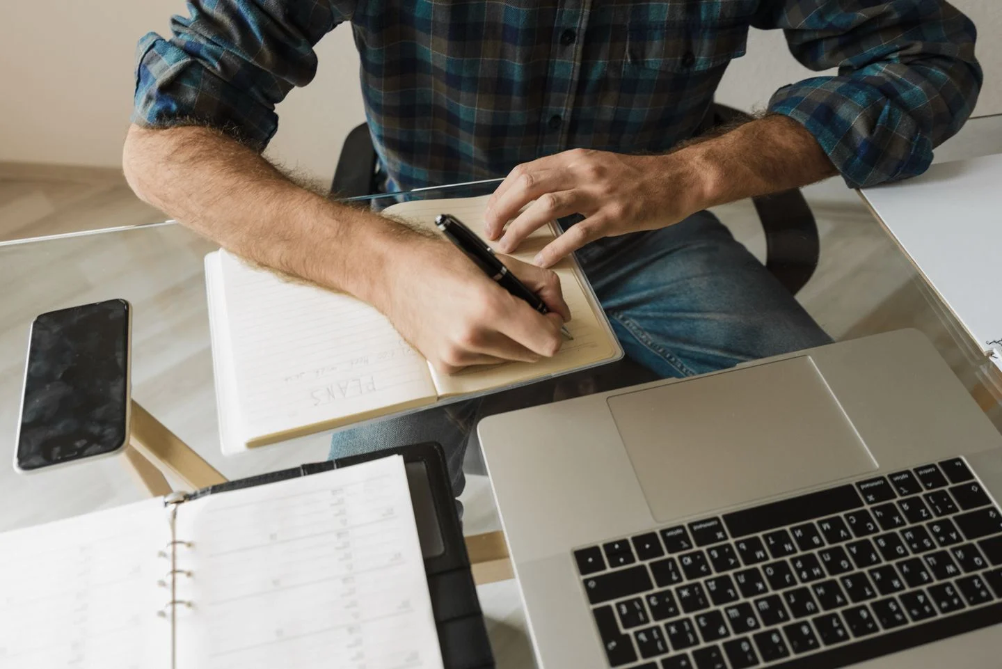 Person writing notes beside a laptop and paper planner at a desk.